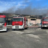 Tre automezzi dei vigili del fuoco rossi parcheggiati davanti a un edificio in fiamme, fumo nero e nuvole sopra, scena di emergenza.