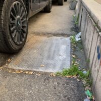 Auto parcheggiata vicino a una rampa di accesso, superficie in cemento e bordo marciapiede, con erba e rifiuti lungo il lato.