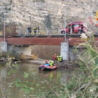 Intervento di soccorso su un ponte: personale in canoa e su barca, mezzo di trasporto rosso e squadra in abiti ad alta visibilità durante operazioni di emergenza.