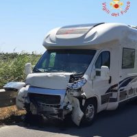 Autocaravan da viaggio bianco danneggiato frontalmente su una strada, con erba sullo sfondo e logo dei Vigili del Fuoco nell’angolo superiore.