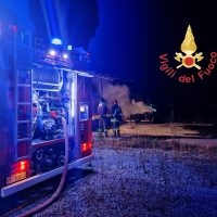 Automezzi dei vigili del fuoco con luci rosse e blu lampeggianti, intervento notturno in campo aperto, fumo e personale all’opera.