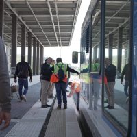 Gruppo di viaggiatori in attesa su una passerella pedonale coperta, con zaini colorati e divise tra vetro e struttura metallica.