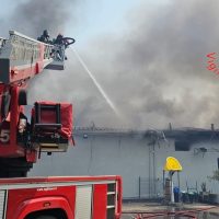 Vigili del fuoco al lavoro, autobotte rossa in primo piano e getto d’acqua su un incendio in edificio industriale, fumo grigio lattiginoso sullo sfondo.
