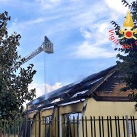 Casa colonica con tetto parzialmente danneggiato e tegole spostate; attrezzi e recinzione visibili, cielo azzurro.