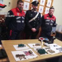 Presso tavolo con oggetti sequestrati: ordigni, moschetti e strumenti, tra agenti in divisa e militari durante un’operazione di polizia locale