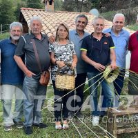 Gruppo di persone anziane all’aperto, sorridenti, in un contesto rurale con palme e una casa in pietra sullo sfondo.