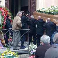 Gruppo di persone in lutto all'ingresso di una chiesa, con corone e fiori, discutono o osservano durante una cerimonia di funerale.