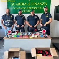 Quattro uomini della Guardia di Finanza in piedi dietro una scrivania con strumenti e involucri, sfondo verde con testi ufficiali, oggetti e scatoloni sul tavolo.