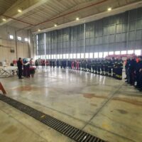 Gruppo di persone in cerimonia all'interno di un grande capannone, in fila lungo i lati, con movimento centrale tra file di addetti.