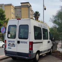 Furgone bianco sul bordo strada, dietro palazzi urbani, segnali stradali e alberi. Lato laterale mostra porte e finestrini, stile urbano.