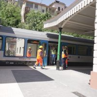 Stazione ferroviaria con trenino elegante blu, operai in alta visibilità arancione sull'atrio, tettoia bianca e edifici verdi sullo sfondo.