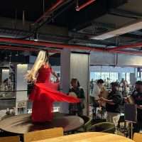 Donna in abito rosso danza su tavolo in un caffè affollato, con luci soffuse e persone sedute al tavolo sullo sfondo.