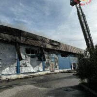 Edificio industriale fatiscente con muri graffiati e porte blu, bastoni di metallo e gru in alto, su strada asfaltata.