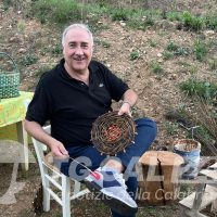 Uomo sorridente mentre intreccia cesti di vimla all'aperto, con tavolino verde e campagna sullo sfondo.