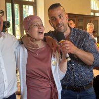 Due persone sorridenti cantano insieme; un uomo tiene un microfono mentre è circondato da amici in un ambiente luminoso e festoso.