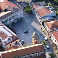 Veduta aerea di una piazza di paese con stradine, edifici colorati, auto parcheggiate e una statua al centro, atmosfera rurale tardo-popolare.