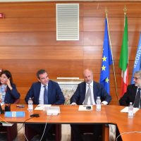 Foto di una riunione formale: quattro uomini seduti a un tavolo in legno, con bandiere europee e italiane sullo sfondo.
