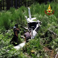 Scena di soccorso in mezzo al verde: elicottero inclinato tra cespugli, soccorritori al lavoro vicino al morente macchinario.