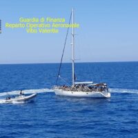 Motovedetta della Guardia di Finanza all’ancora nel mare azzurro, piccolo gommone a fianco, testo giallo in alto