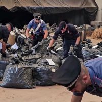 Gruppo di uomini che raccolgono e spostano sacchi neri in un'area disordinata, indossando caschi e pettorine, in un contesto di lavoro pesante.