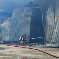 Vigile del fuoco vicino a una barriera di metallo arrugginita, spruzza acqua su un incendio, con getto rosso di gomma e montagne sullo sfondo.