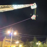 Cantiere con gru a mezzanotte, braccia articolate e funi illuminate, sfondo urbano con lampioni e edifici.