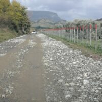 Strada sterrata con ghiaioni, fango e pali di recinzione in primo piano, paesaggio collinare sullo sfondo, cielo nuvoloso.