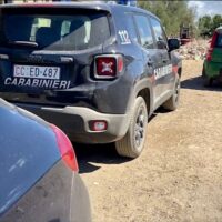 Auto dei Carabinieri in primo piano, SUV grigio scuro con targa CC e scritte sui fianchi, parcheggiata su terreno di ghiaia.