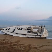 Barca a vela bianca arenata sulla riva sabbiosa, mare calmo all’orizzonte e cielo sereno al tramonto.