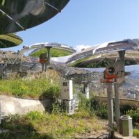 Panoramica di ombrelloni solari all'aperto su terrazza con vista città, cielo sereno e colture in primo piano.