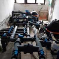 Impianto di tubazioni e valvole nere con raccordi blu, all’interno di una stanza, visibili tubazioni, pompe e serbatoi.