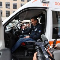 Medic in ambulanza seduto al posto guida, intervista video da parte di media; persona sorridente durante una procedura di soccorso in città.