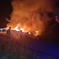 Incendio acceso su un veicolo abbandonato di notte, fiamme arancioni e fumo alto; una persona osserva da distanza sulla strada.