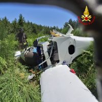 Incidente aereo: fusoliera di un piccolo aereo ribaltata tra erba alta, parte del muso danneggiata e operatori sullo sfondo.