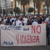 Rete di persone in protesta con cartellone: NO VIOLENZA e CROTONE dice, scena all'aperto in piazza, palme e edifici sullo sfondo.