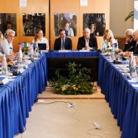 Sala riunioni allargata con tavolo a forma di U, presenti professionisti in abiti formali, elementi decorativi e piante centrali.
