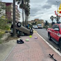 Automobile capovolta su marciapiede, oggetti sparsi a terra, veicoli in strada con presenza di soccorso e palme sullo sfondo, strada urbana.