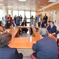 Ufficio affollato: riunione formale in sala conferenze con partecipanti seduti attorno a tavolo, alcuni in rispettosa standing, discussione animata.