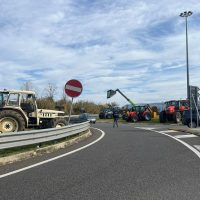 Immagine di una rotonda stradale con veicoli agricoli parcheggiati sul lato sinistro e una gru allestita al centro, cielo sereno.