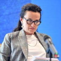 Donna in giacca grigia legge documenti durante un incontro, sfondo blu. L'immagine è professionale e formale.