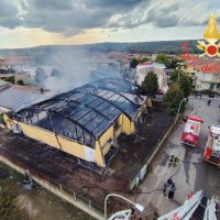 Incendio in un piccolo capannone giallo: fumo che si alza, vigili del fuoco e camion presenti, quartiere urbano circostante.