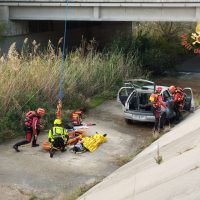 Soccorritori trasportano un ferito in barella vicino a un’auto incidentata; intervento in un’area di cortile con vegetazione e presenza di gente.