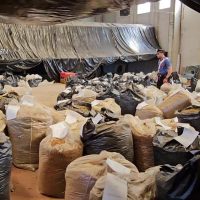 Stagno di sacchi beige riempiti di materiali, in magazzino interno con teli e scatoloni, figura umana in lontananza tra gli ingombranti.