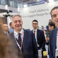 Uomo in abito elegante sorride tra altri partecipanti a una fiera, sfondo con banner testuale e conferenza. Attenzione a dettagli professionali.