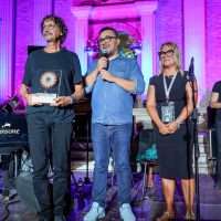 Tre musicisti su palco illuminato con luci rosa: due uomini parlano, una donna ascolta, atmosfera vivace e concerto in corso.