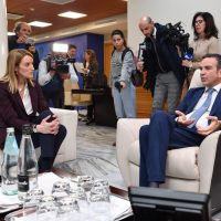 Uomo in smoking e donna in abito seduti su divani, interviene durante conferenza con relatori e media presenti in sala.