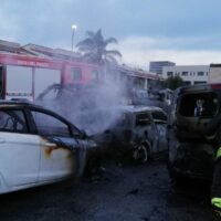 Auto dimearsa annerita e fumante su una strada, soccorritori all’opera tra veicoli gravemente danneggiati e fumo.