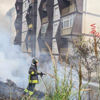 Pompieri in azione davanti a un edificio alto, getto d'acqua e fumo, scenario urbano con vegetazione e segnalazione visiva a destra.