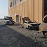 Auto d'epoca parcheggiate lungo una strada acciottolata di fronte a un edificio beige, con fumo e alberi sullo sfondo.