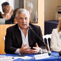 Uomo anziano in giacca scura parla al tavolo durante una conferenza, gesticolando con mani aperte.